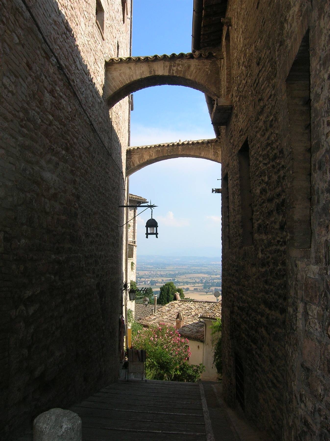 Stone arches between medieval houses in Assisi, sunlit Umbrian valley below