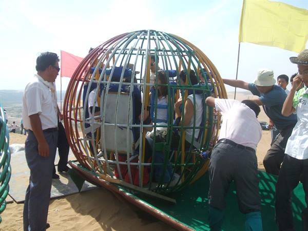Tourists inside a metal ball ride, helpers push it across the sand