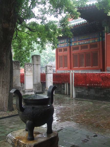 Bronze incense burner in rainy temple courtyard, red prayer tablets and stone stele