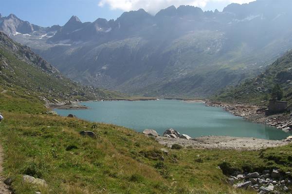 Turquoise alpine lake between meadows and rocky peaks, hiker on trail