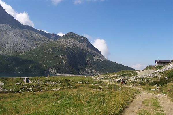 Hikers on grassy mountain trail toward dam and alpine lake among granite rocks