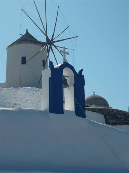 White and blue belfry with cross in front of a windmill in Oia, Santorini