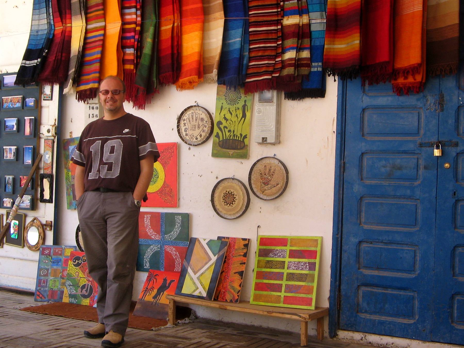 Man smiling in front of colorful rug stall and paintings, blue door