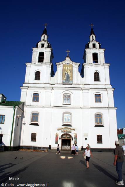 White cathedral with two bell towers in Minsk, tourists walk on the square under blue sky