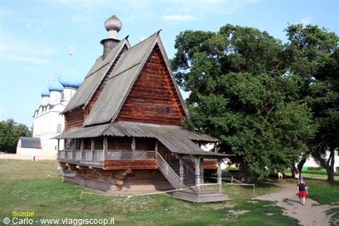 Wooden church in Suzdal, blue kremlin domes behind, a tourist walks by