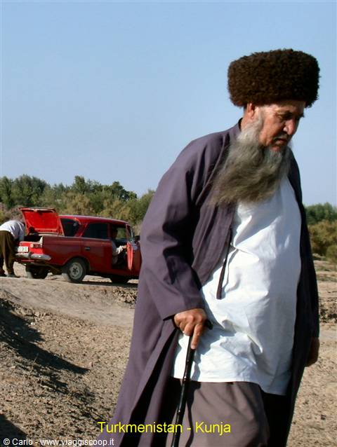 Elderly Turkmen man with long grey beard and fur hat walks with cane, red car behind