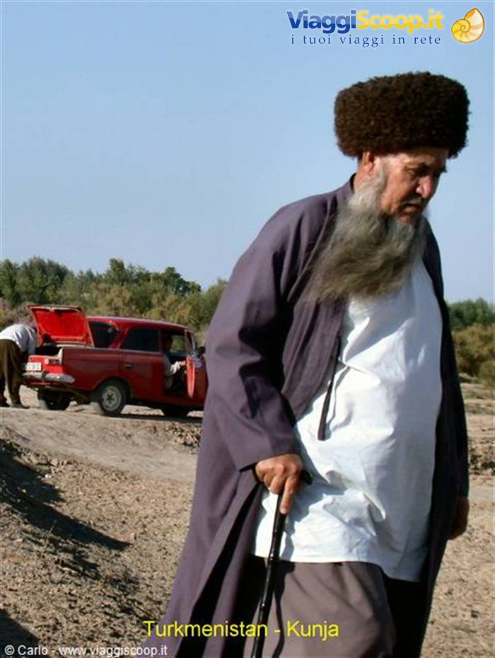 Elderly Turkmen man with long grey beard and fur hat walks with cane, red car behind