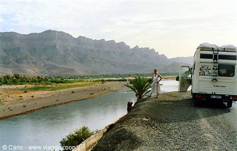 Motorhome parked by a river with palm trees and rocky mountains