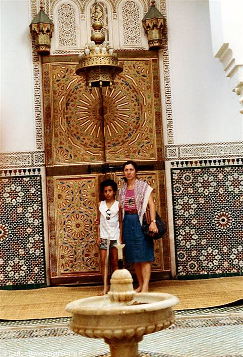 Woman and child before the mosque's carved brass door, zellige tiles, marble fountain