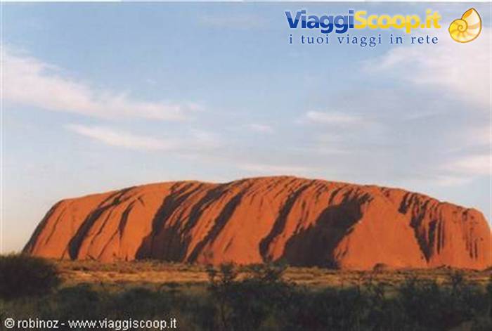 Uluru, red sandstone monolith, rises among shrubs under white clouds