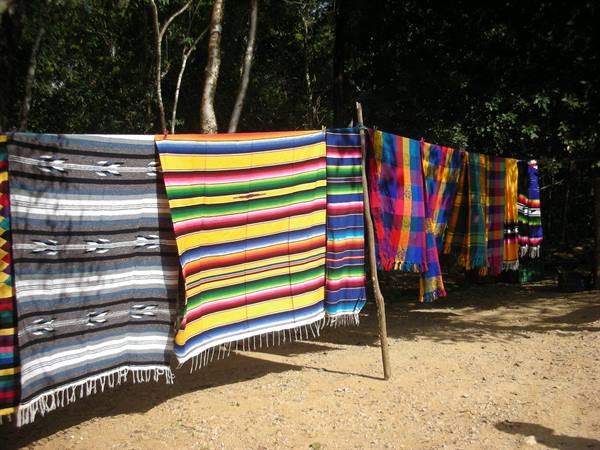 Colorful woven blankets hanging on a wooden branch among trees, red dirt ground