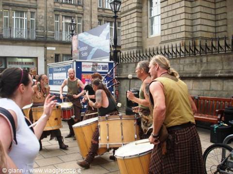 Drummers in kilts playing large drums on a stone street among buildings