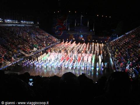 Massed performers in colorful uniforms parade in the dark arena before a packed crowd
