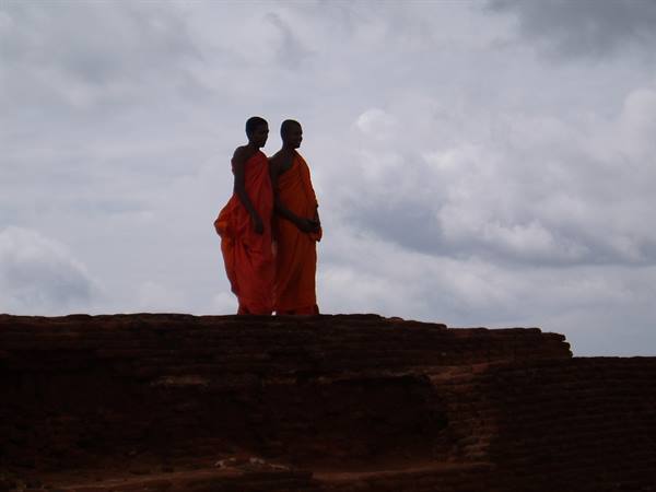 Two monks in orange robes walk on ancient brick ruins under grey clouds