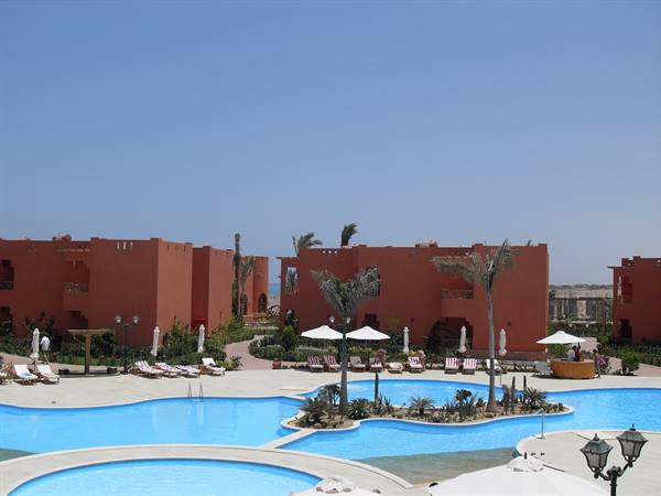 Blue pools among palms and umbrellas, ochre resort buildings, clear sky