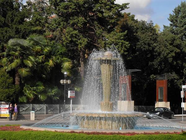 Stone fountain spraying water among palm trees and greenery, pedestrians walking by