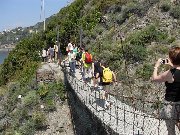 Hikers with backpacks cross a wire-mesh suspension bridge on the Ligurian coast