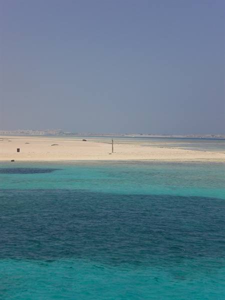 Turquoise waters in front of a pale sandy beach, building in the distance