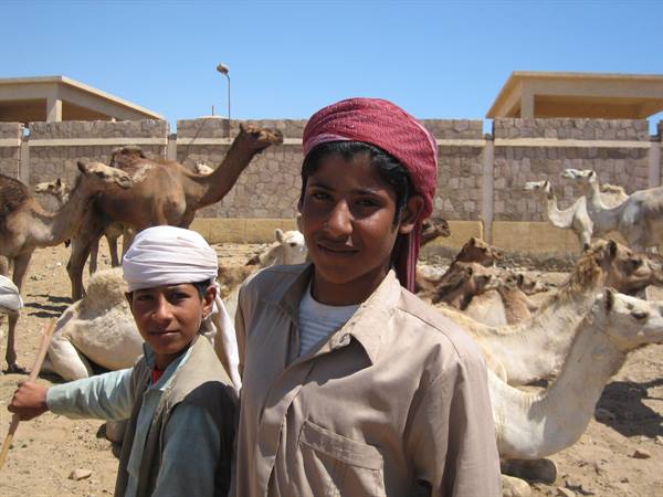 Two smiling boys in headscarves among camels at a sandy market