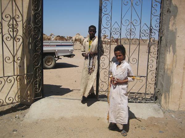 Two boys in white robes by a wrought-iron gate, camels and pickup truck behind