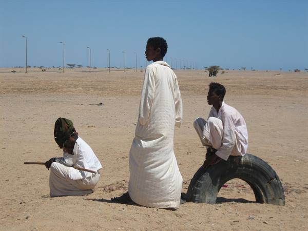 Three boys in white robes on desert sand, one sitting on a tire