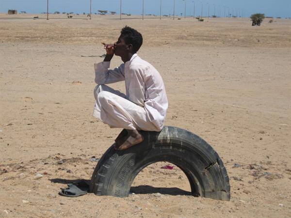 Man in white robe sitting on a large tire in the sandy desert