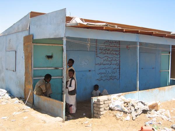 Children in front of a blue metal shack with Arabic writing, sand and harsh sunlight