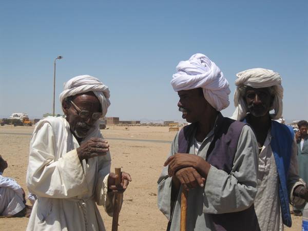 Three men in white turbans holding wooden canes in an arid desert