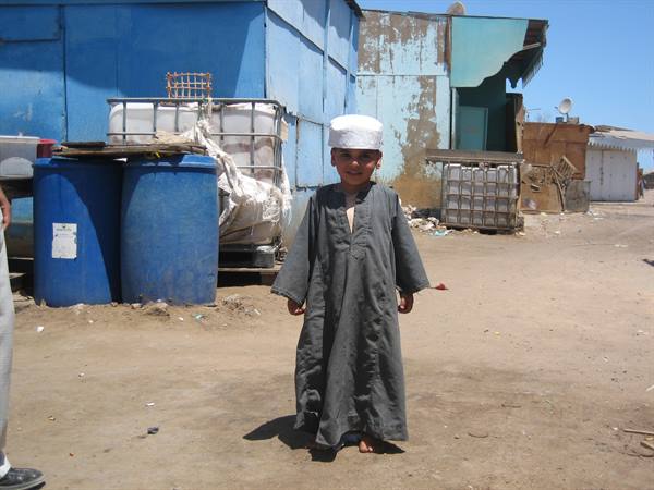 Smiling boy in grey galabeya and white cap on sandy street among blue barrels