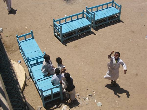 Children playing near blue wooden benches on sandy ground