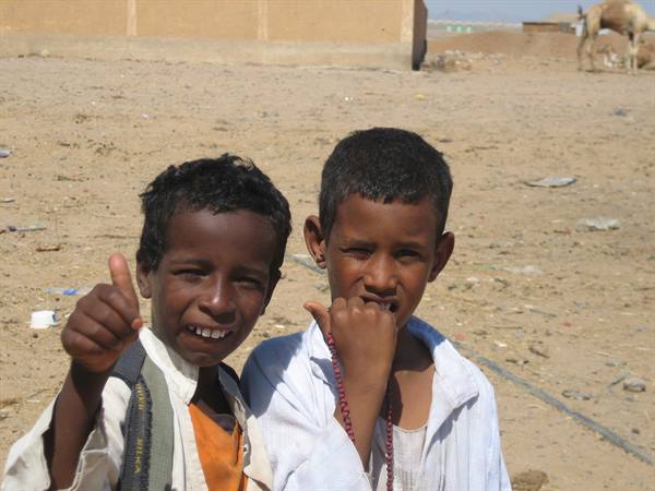 Two smiling boys giving thumbs up in a desert village with a camel