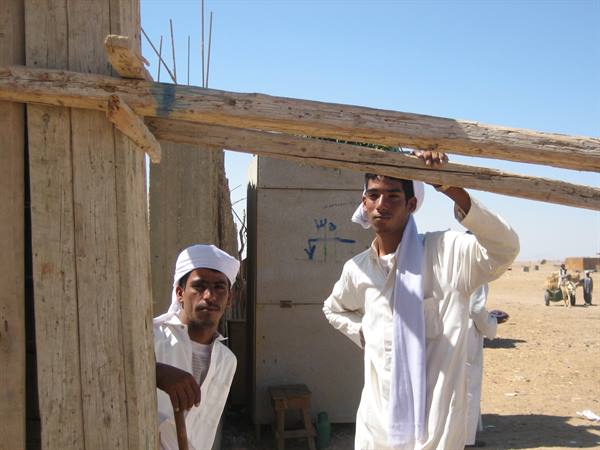Two young men in white galabeyas and turbans lean on wooden beams in the desert