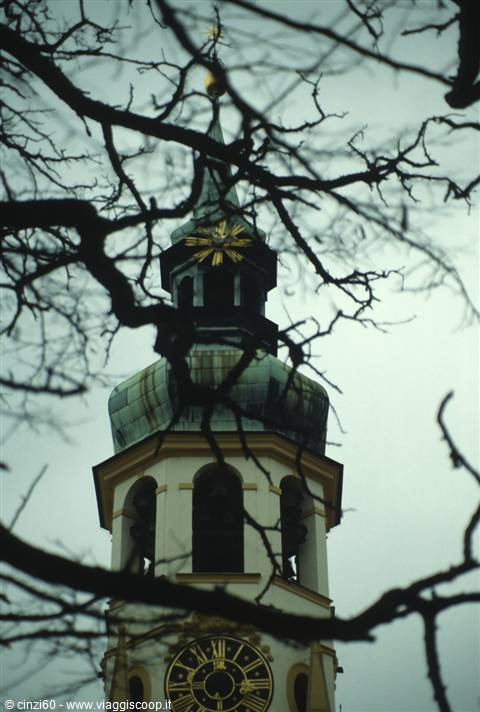 Baroque church tower with green copper dome and golden clock behind bare branches