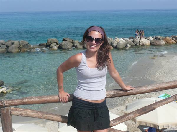 Smiling woman in sunglasses leaning on a wooden railing by a rocky turquoise beach