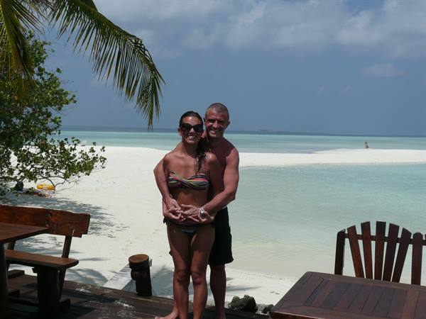Couple embracing on white sand beach, turquoise water, palm tree and wooden furniture