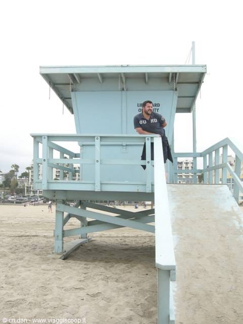 Man leaning on the railing of a light blue lifeguard tower on the sand