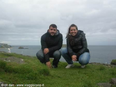 Two people crouching on grassy cliffs overlooking the grey ocean under cloudy skies