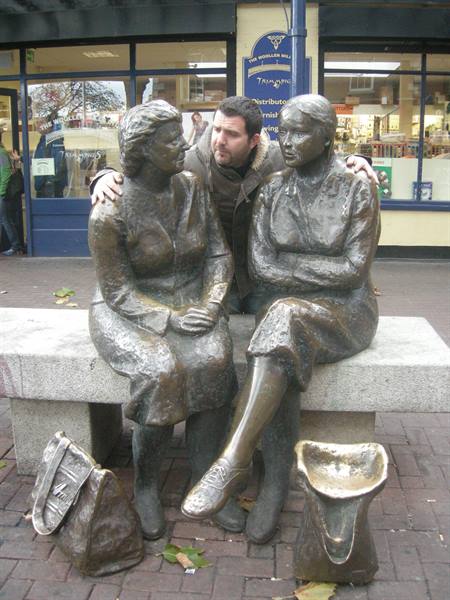 A man embraces two bronze statues of seated women from behind on a bench