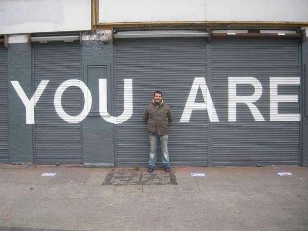 Smiling man in green jacket leaning on grey shutters painted YOU ARE