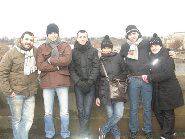 Six smiling friends in winter clothes leaning on a stone wall, river and buildings behind