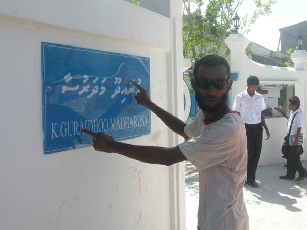 Man in sunglasses pointing at the Guraidhoo Madharusa school sign