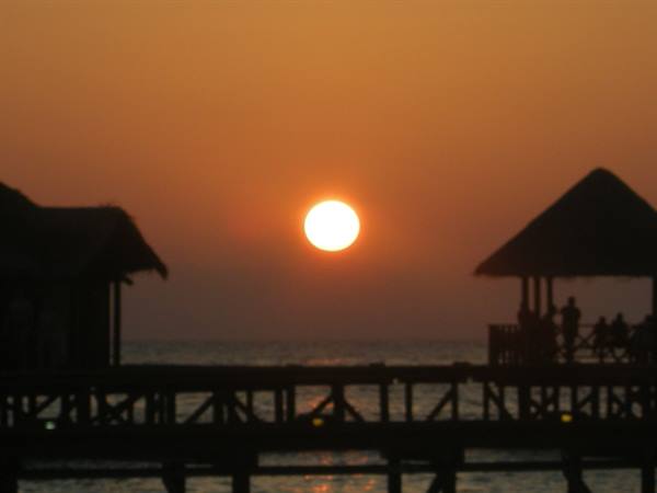 Low sun over the ocean between wooden stilt huts with thatched roofs