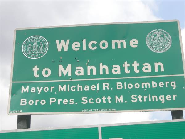 Green highway sign reading Welcome to Manhattan, with official seals, against cloudy sky