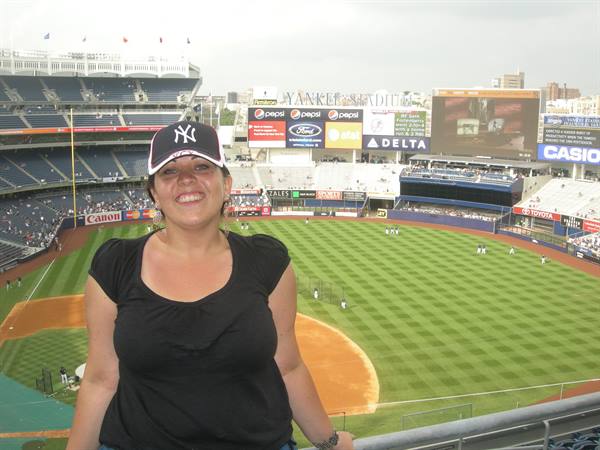 Woman in Yankees cap smiles at Yankee Stadium, baseball field daylight