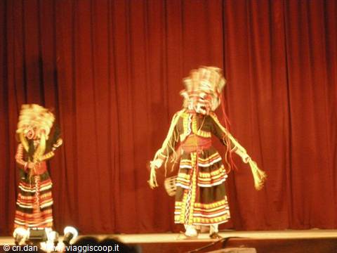 Masked traditional dancers in colorful striped skirts perform before a red curtain
