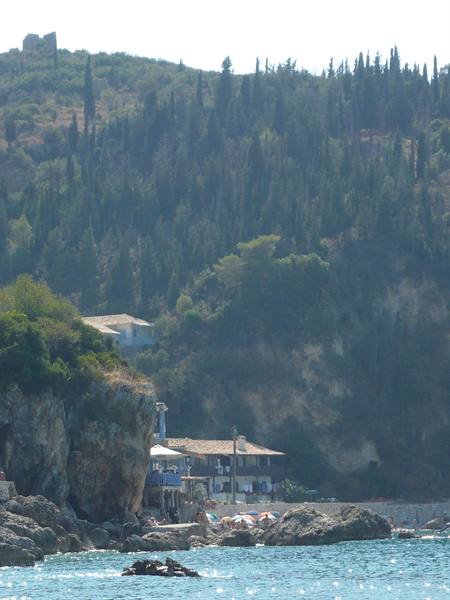 Houses and beach umbrellas on rocky shore below cypress-covered hill, Corfu