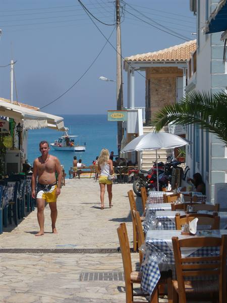 Stone lane with checkered tables, people walking to blue sea with boat