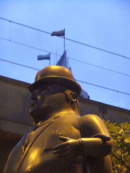 Bronze Botero statue wearing helmet with briefcase, flags against blue sky