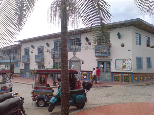 Parque Municipal Guatape: white building with wooden balconies, colorful tuk-tuks and palms