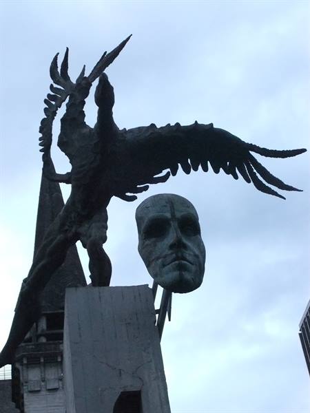 Bronze statue with large mask and winged figure against cloudy sky